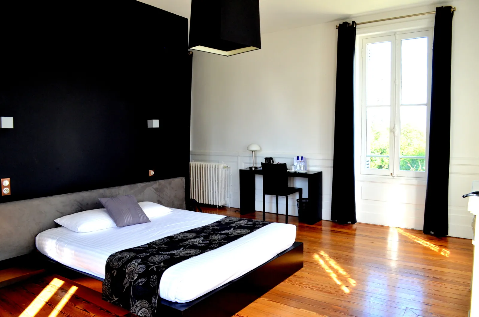 Chambre moderne au Château La Chèze à Bordeaux avec lit king-size aux draps blancs, mur noir caractéristique, parquet en bois, bureau noir, portes-fenêtres donnant sur le jardin et mobilier contemporain.