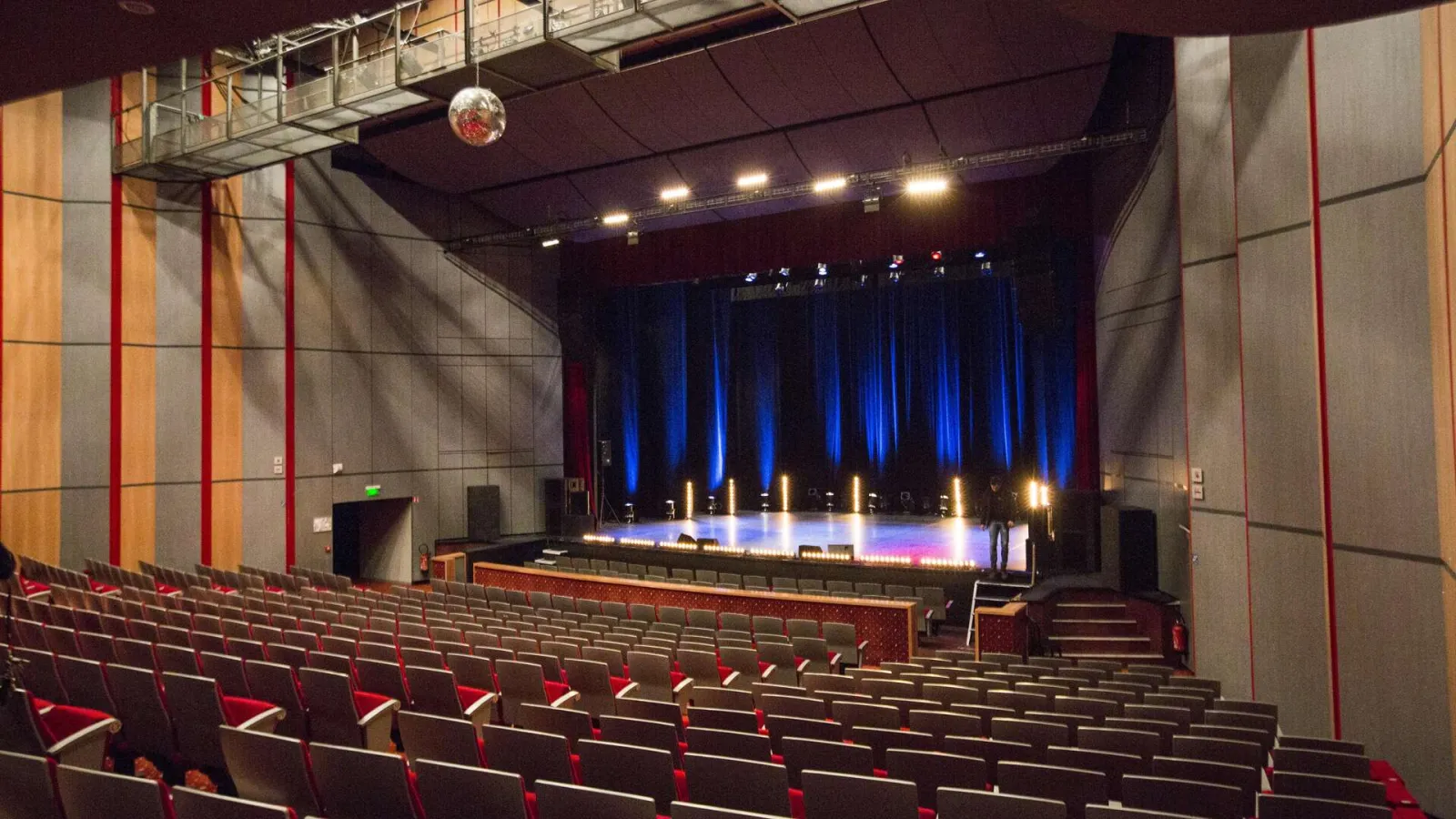 Intérieur d'une salle de théâtre moderne à Bordeaux avec des sièges rouges en amphithéâtre, un plafond sombre, des murs géométriques beige et rouge, et une scène illuminée par des projecteurs bleus et blancs.