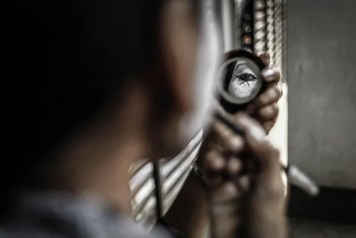 Gros plan d'une main tenant un miroir rond reflétant un visage, avec un arrière-plan flou et des barreaux visibles, dans une atmosphère urbaine sombre.
