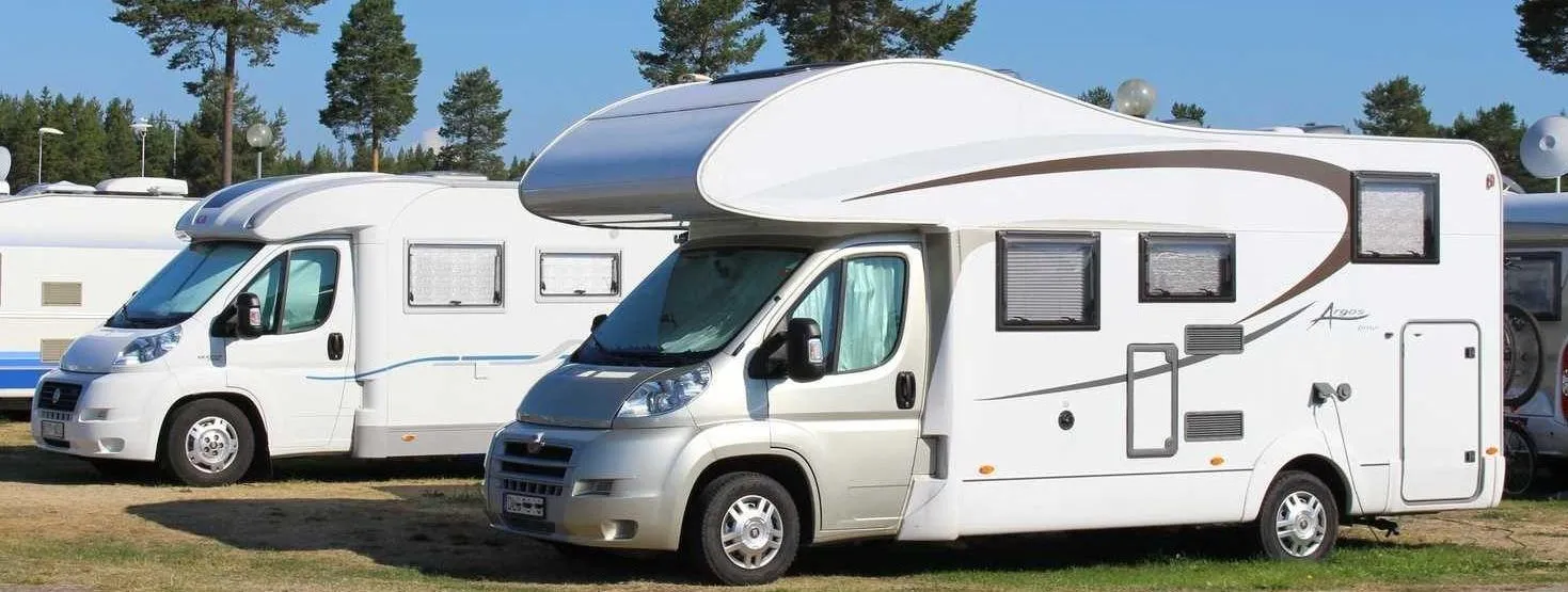 Trois camping-cars de différentes tailles stationnés côte à côte sur une aire de parking, avec des arbres en arrière-plan et un ciel dégagé.