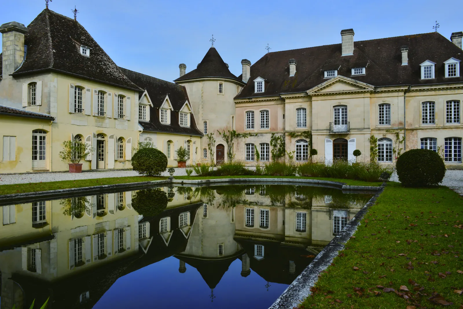 Château Bouscaut à Bordeaux, élégante demeure classique avec façade en pierre de couleur crème, toits ardoise et mansardes, reflétée dans un bassin d'eau rectangulaire, entourée de jardins à la française avec buis taillés.