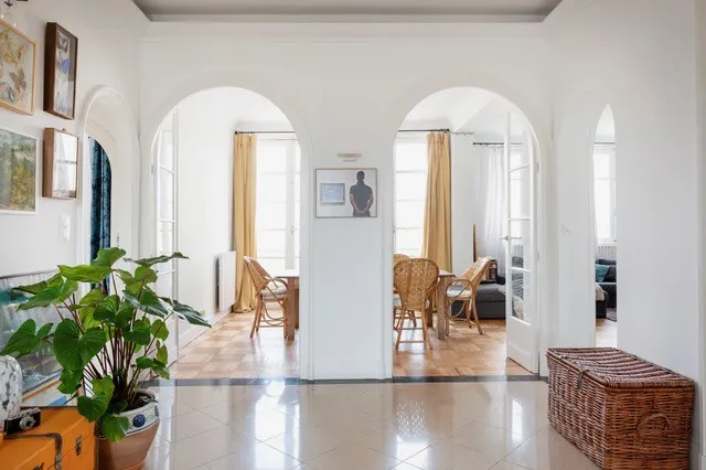 Intérieur lumineux d'un appartement bordelais avec hall central aux arches blanches, sol en terrazzo brillant, plante verte en pot orange, mobilier en rotin et portes ouvertes sur les pièces adjacentes