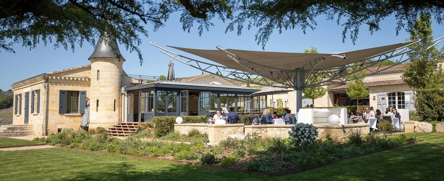 L'Atelier de Candale à Bordeaux : bâtiment en pierre de taille avec tour, extension vitrée contemporaine, terrasse ombragée et clients assis sous voile d'ombrage