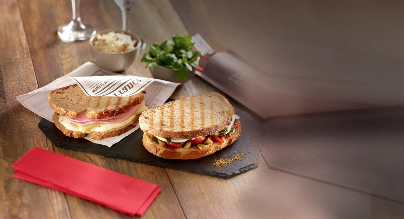 Deux sandwichs grillés sur pain de mie servis sur ardoise noire à l'Ibis Kitchen Lounge de Bordeaux, accompagnés d'une salade, d'une serviette rouge et de verres de vin