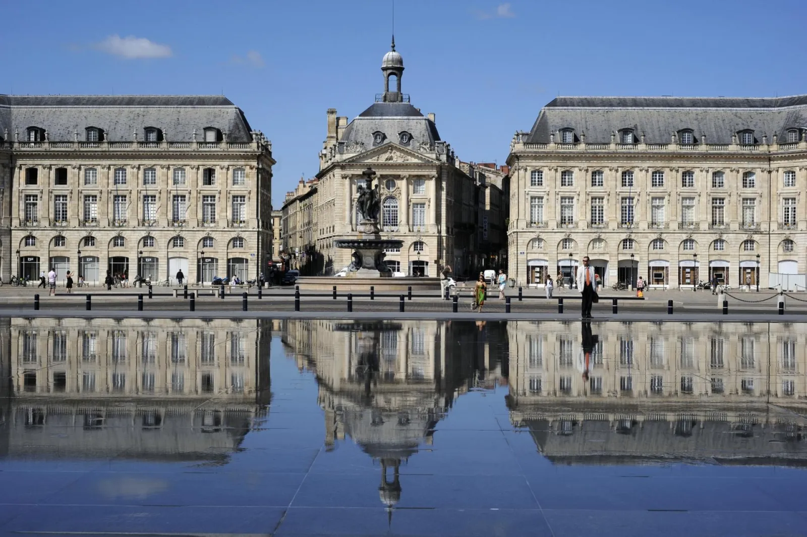 Place de la Bourse à Bordeaux avec ses façades classiques du XVIIIe siècle reflétées dans un miroir d'eau, statue équestre au centre, ciel bleu dégagé.