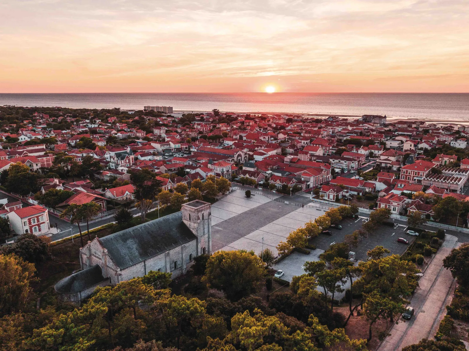 Vue aérienne au coucher de soleil de Bordeaux avec la Basilique Notre-Dame-de-la-fin-des-Terres au premier plan, entourée de maisons aux toits rouges et de verdure, dominant la Gironde.