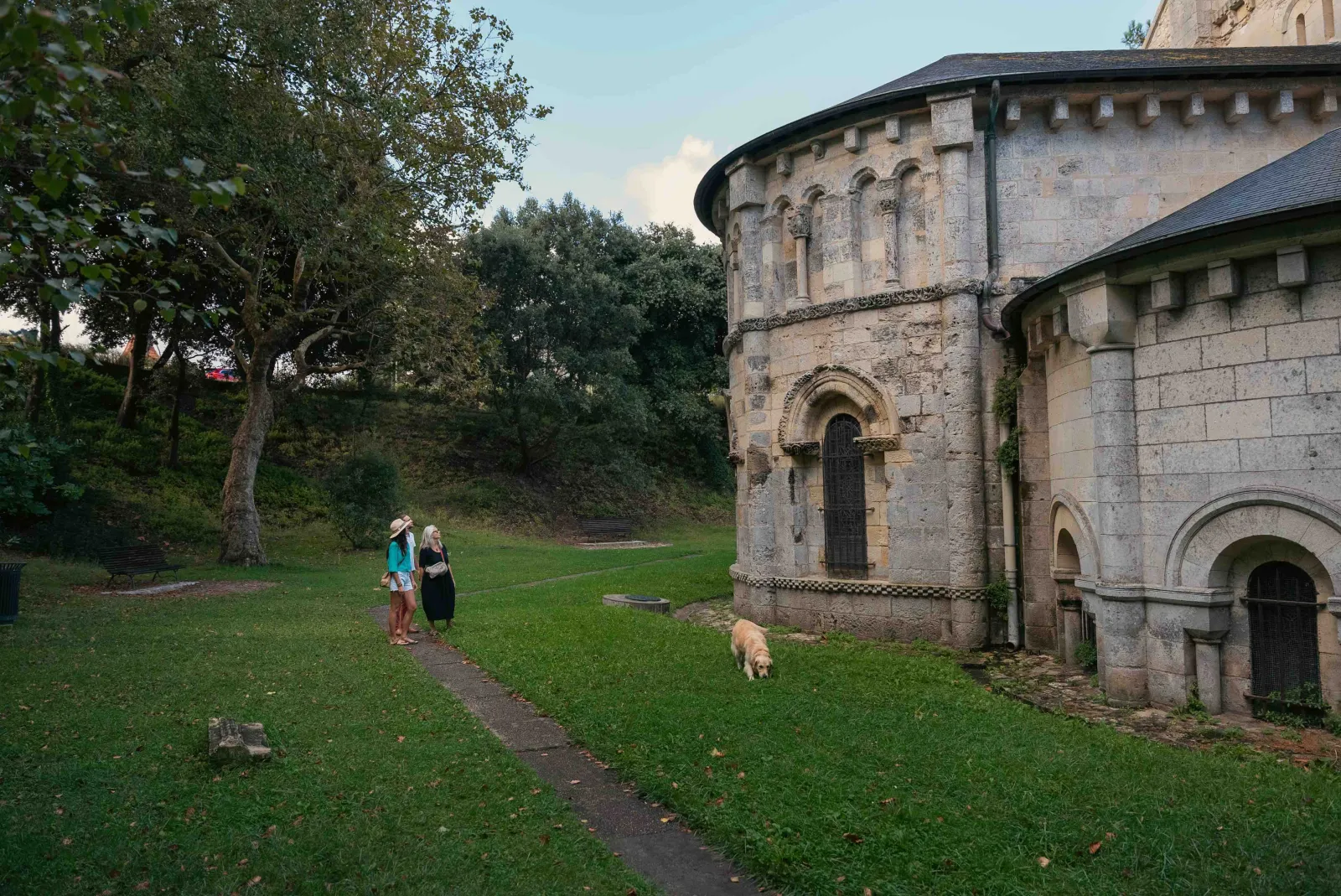 La basilique Notre-Dame-de-la-fin-des-Terres à Bordeaux avec ses murs en pierre, ses arcs romans et sa tour ronde, entourée d'une pelouse verte où deux visiteurs se promènent avec un chien.