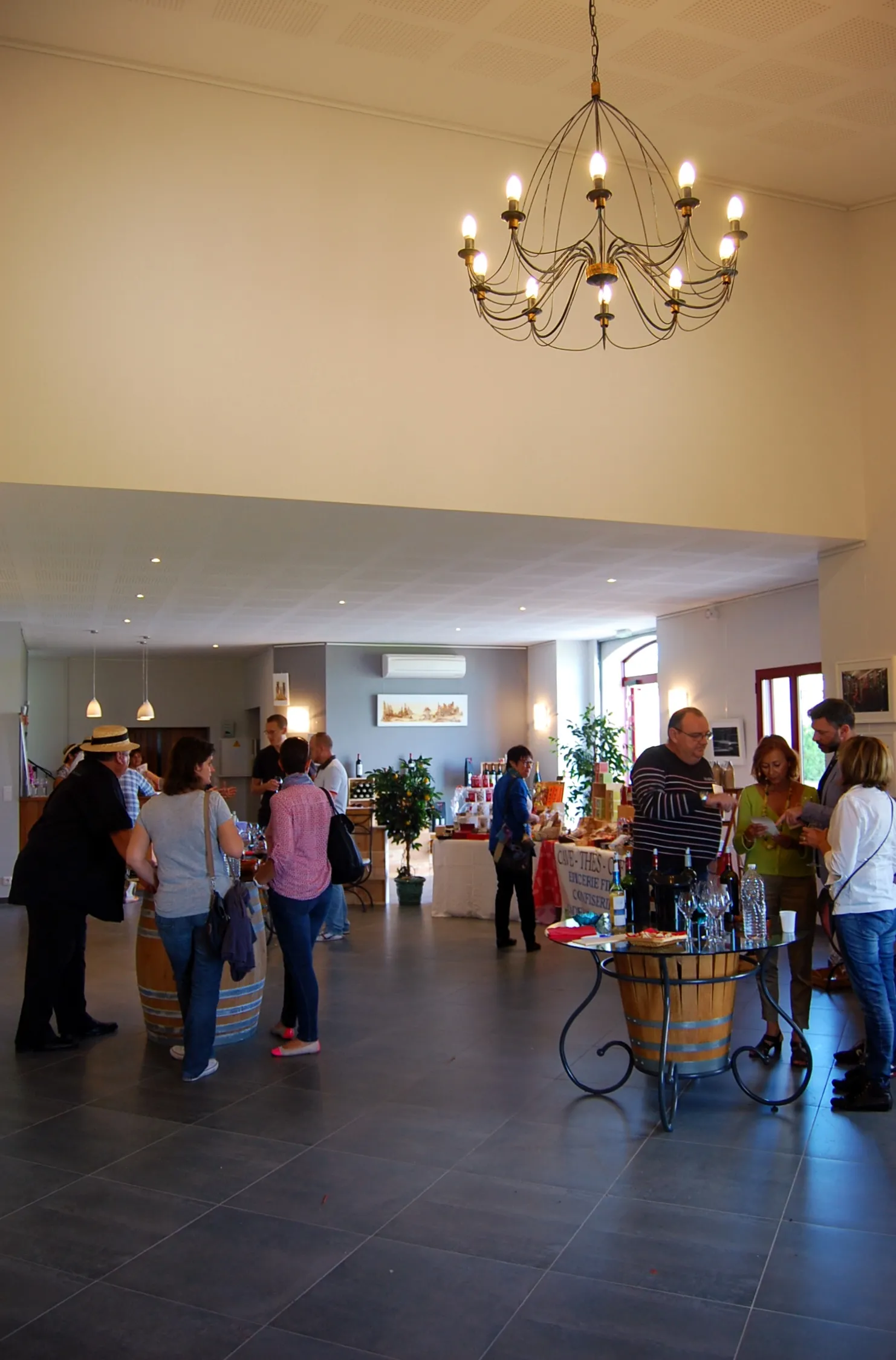 Intérieur spacieux du Château Pichon Bellevue à Bordeaux avec un groupe de visiteurs dispersés dans une salle aux murs beige et gris, éclairée par un lustre en fer forgé ornementé et des spots encastrés, avec un comptoir de dégustation au premier plan.