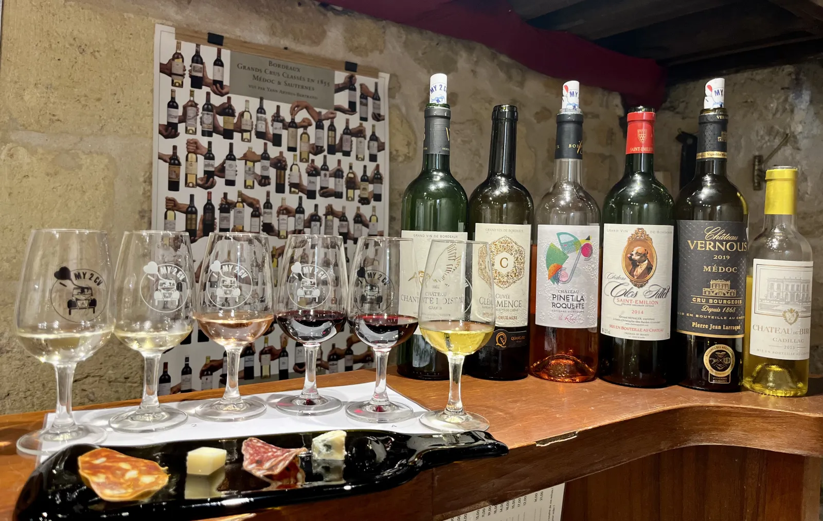 Dégustation de vins de Bordeaux avec six verres contenant différents vins, six bouteilles de Bordeaux et une assiette de charcuterie et fromage, sur fond de poster des Grands Crus de Bordeaux.