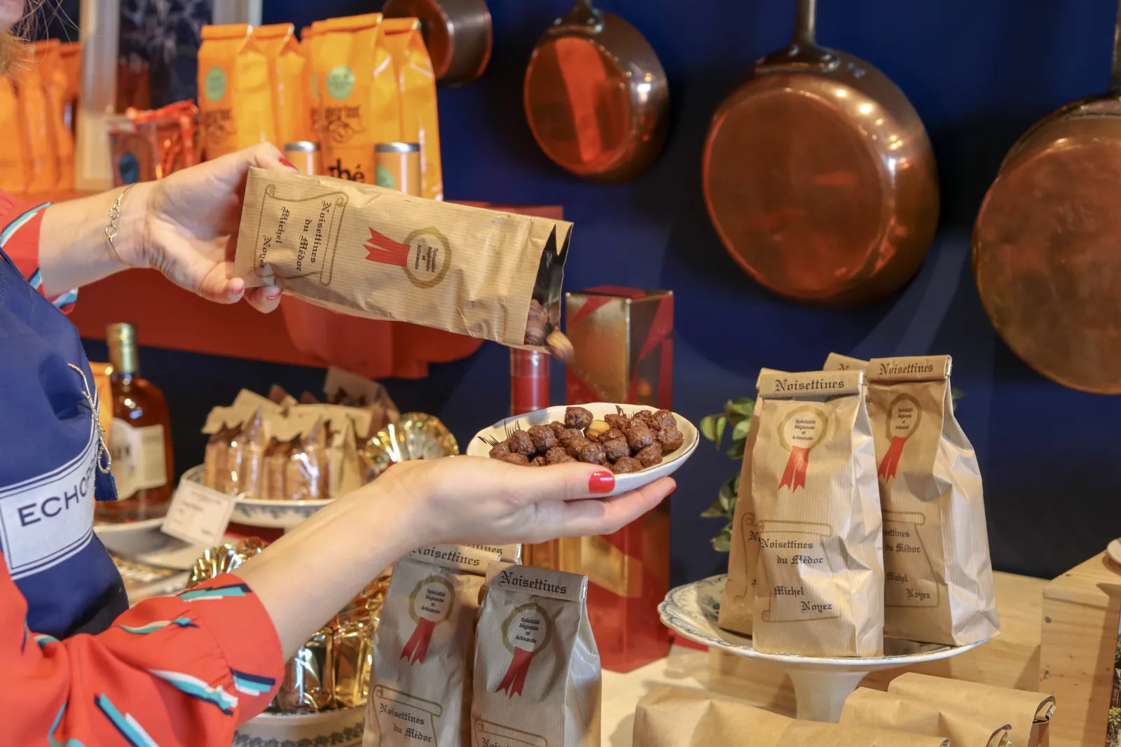 Présentation de noisettes de Bordeaux dans une échoppe, avec une personne tenant un sachet de produit et une assiette de noisettes grillées, entourée de bouteilles et d'emballages artisanaux.