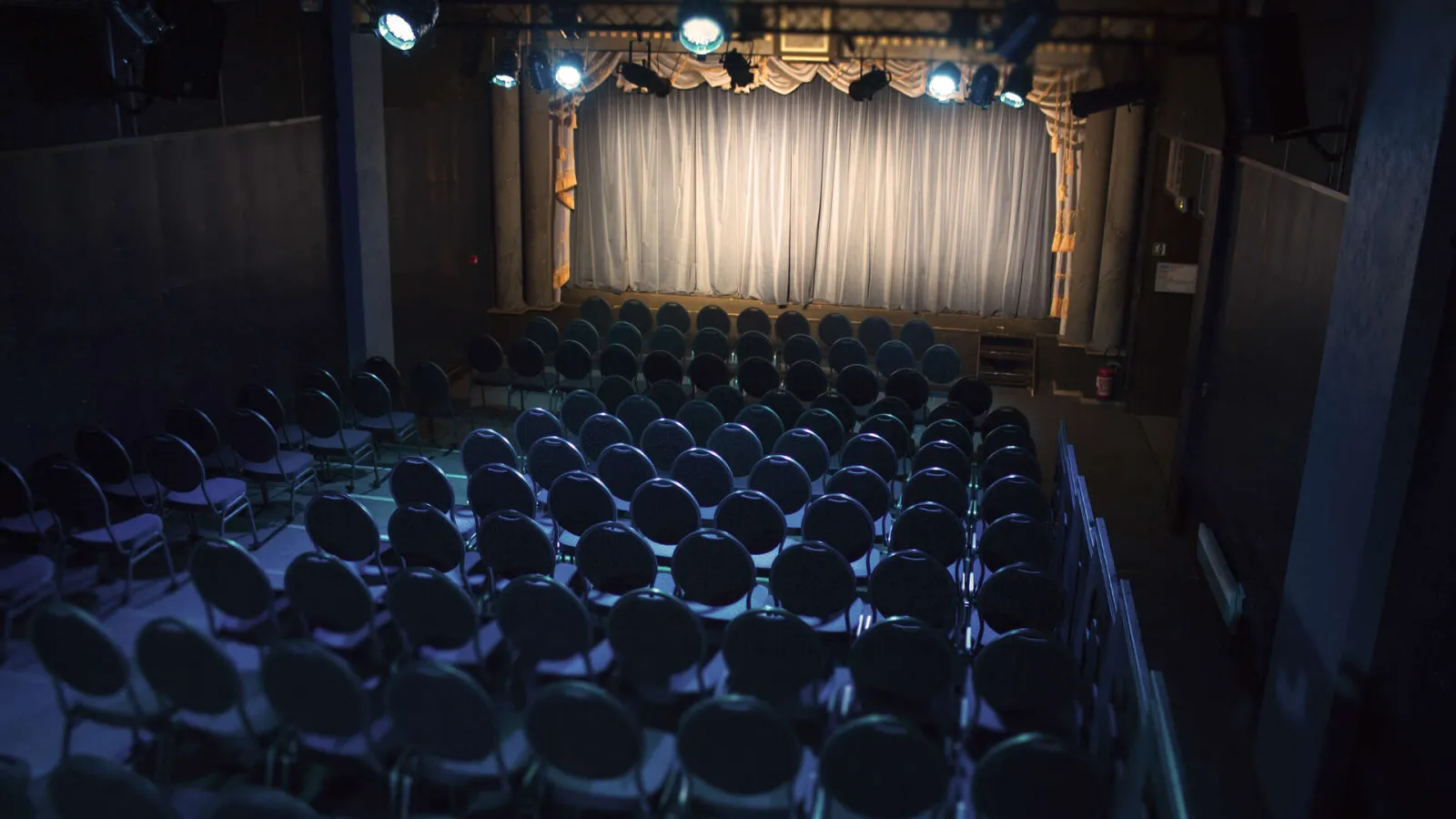 Salle de théâtre vide avec rangées de fauteuils noirs face à une scène illuminée avec rideau blanc et ornements dorés, à Bordeaux.