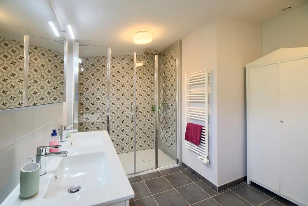 Salle de bain moderne avec double vasque blanche, miroir, douche à l'italienne avec paroi vitrée et carrelage géométrique, radiateur sèche-serviettes blanc et accessoires de toilette