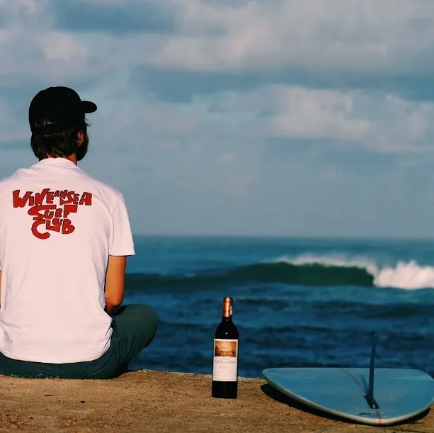Un homme assis sur le sable face à l'océan, portant un t-shirt blanc avec un logo rouge, une bouteille de vin de Bordeaux posée à côté de lui et une planche de surf bleue visible sur la plage.