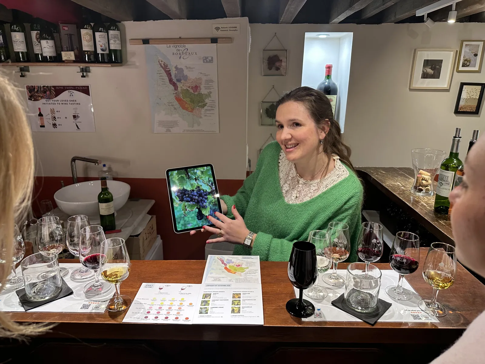 Une femme souriante en pull vert présente une tablette affichant des raisins de raisin lors d'une dégustation de vin à Bordeaux, entourée de verres de vin et de documents informatifs sur les cépages.