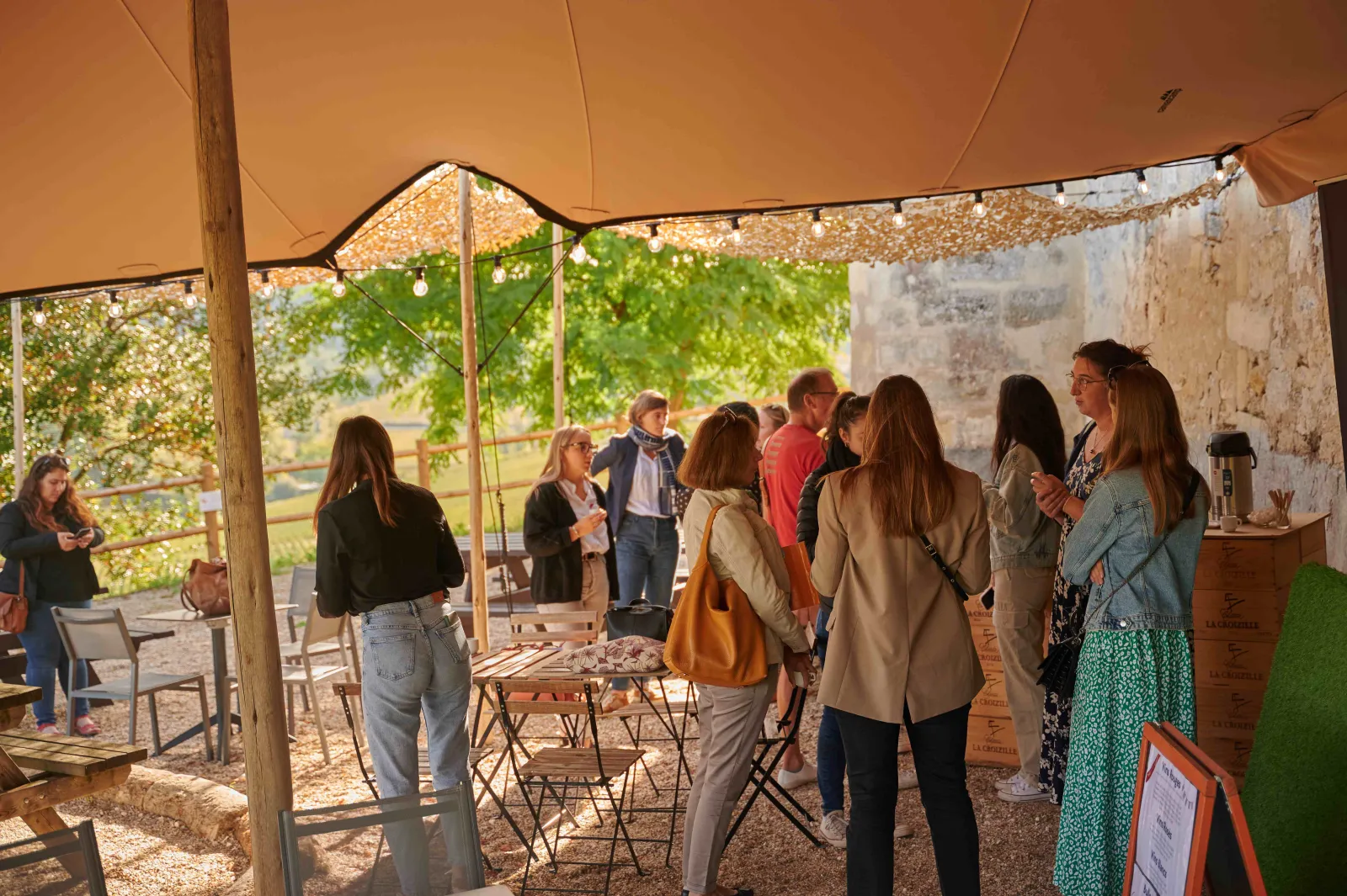 Groupe de visiteurs dans une tente aménagée au bar à vin du Château Tour Baladoz à Bordeaux, discutant et dégustant du vin sous une toile orange.