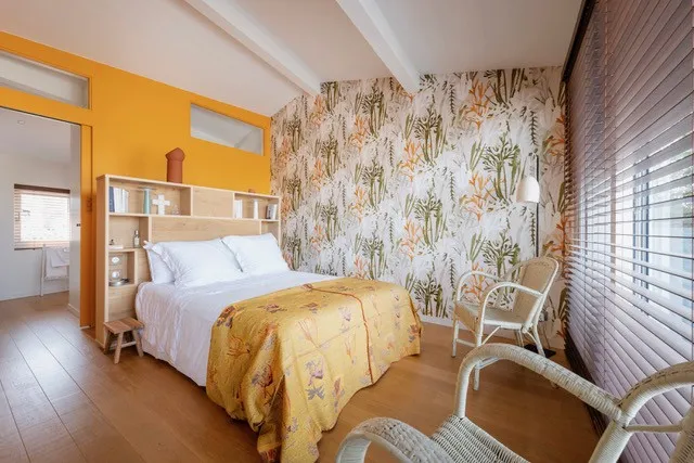 Chambre moderne à Bordeaux avec mur jaune, papier peint floral, lit blanc avec édredon jaune, mobilier en bois clair et chaises blanches près des baies vitrées.