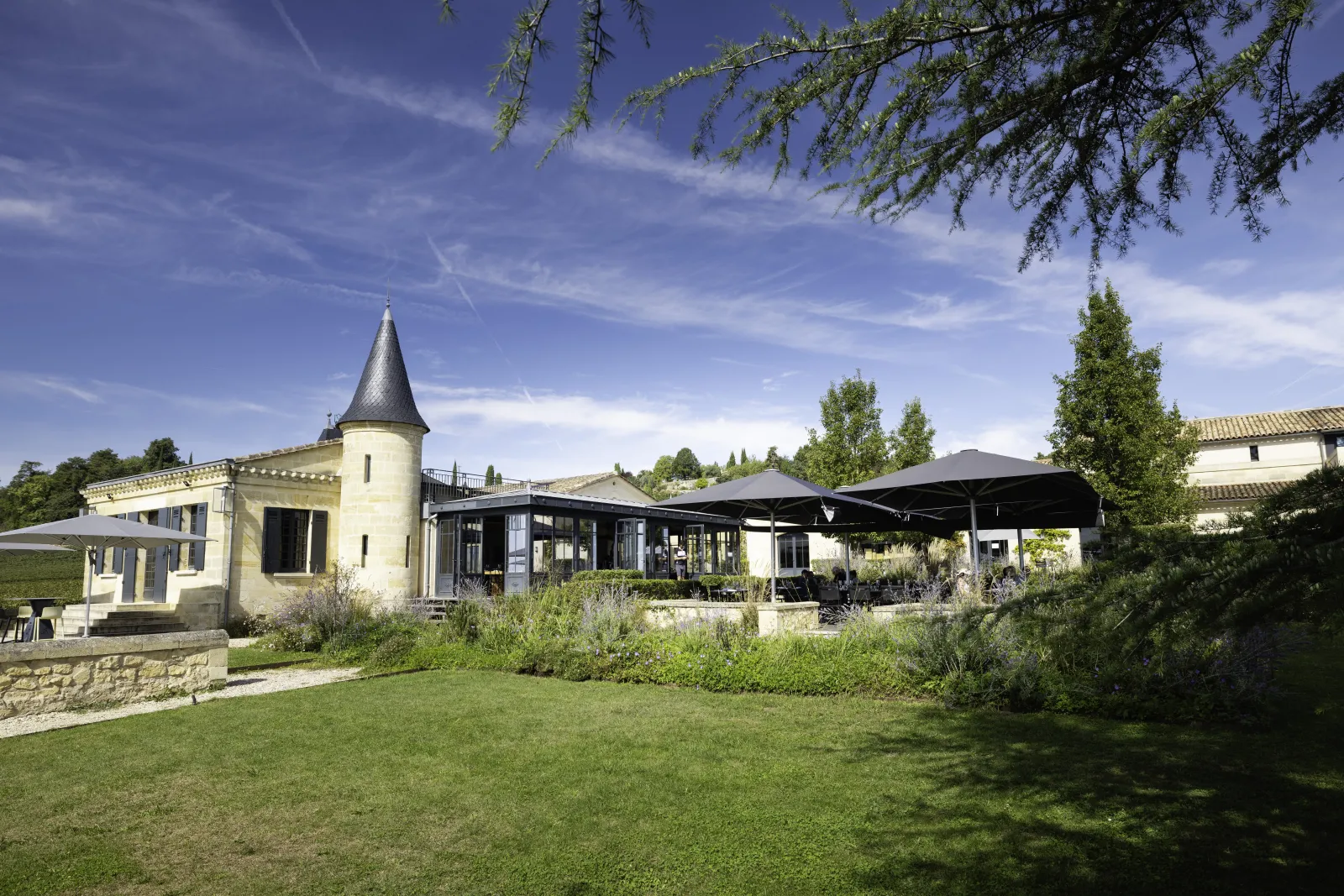 L'Atelier de Candale à Bordeaux : ensemble architectural avec tour pointue, bâtiments modernes aux façades vitrées, terrasse ombragée et pelouse verte, sous un ciel bleu.