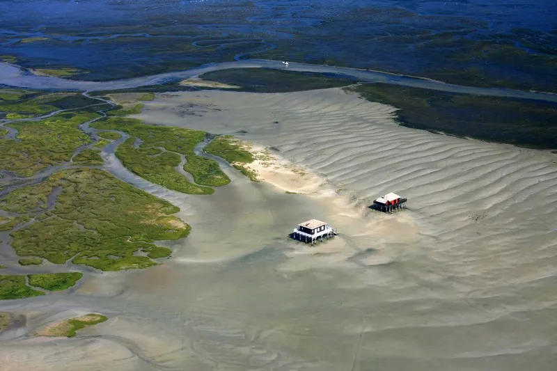 Vue aérienne de l'Île aux Oiseaux près de Bordeaux avec deux cabanes tchanquées traditionnelles sur des bancs de sable, entourées de marais et de cours d'eau sinueux.