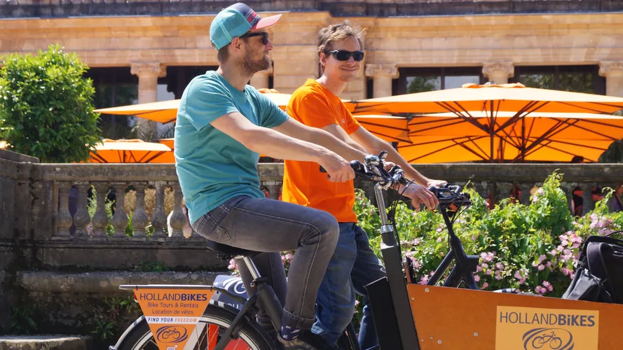Deux cyclistes sur des vélos de location Holland Bikes à Bordeaux, devant des parasols orange et des fleurs, lors d'une visite guidée.