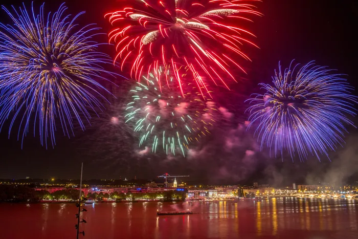 Feu d'artifice du 14 juillet à Bordeaux avec des fusées rouges, vertes et bleues explosant au-dessus de la Garonne, illuminant la ville et ses reflets dans l'eau.