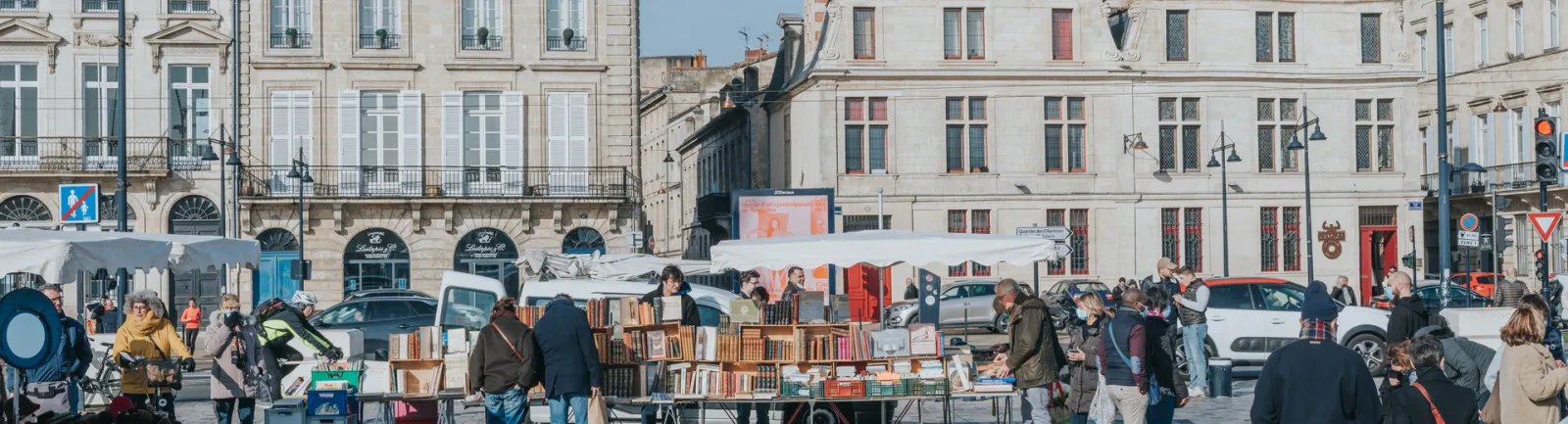 Marché du quai des Chartrons à Bordeaux avec des étals de livres d'occasion sous des tentes blanches, des bâtiments haussmanniens en arrière-plan et des visiteurs flânant entre les stands par une journée ensoleillée.