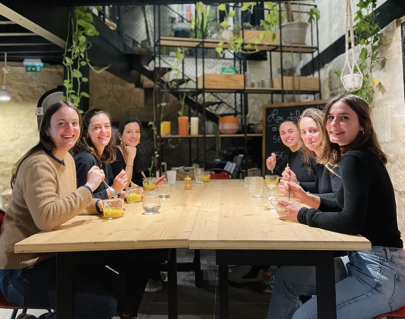 Groupe de sept jeunes femmes souriantes assis autour d'une table en bois dans un restaurant moderne de Bordeaux, avec des verres de boisson et des plantes vertes en arrière-plan.