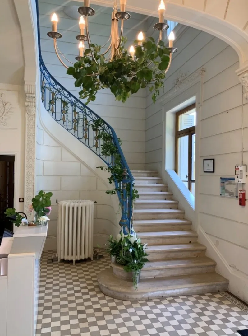 Escalier élégant du Château La Chèze à Bordeaux avec rampe bleue décorée de lierre, chandelier suspendu orné de feuillage, carrelage en damier noir et blanc, et portes en arche blanche