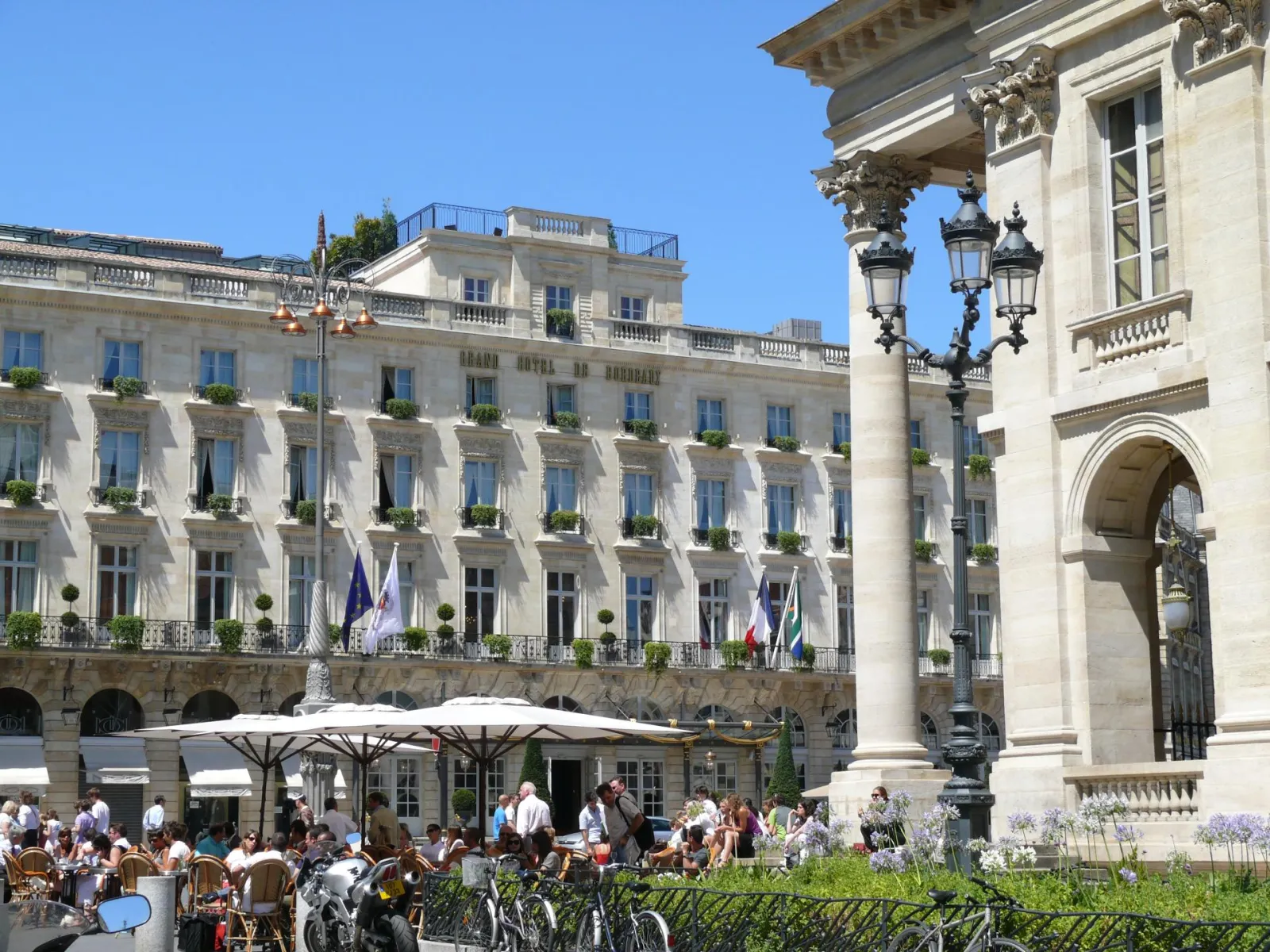 Façade élégante de l'Intercontinental Grand Hôtel de Bordeaux avec ses colonnes corinthiennes, ses balcons fleuris et sa terrasse animée en premier plan sous un ciel bleu.
