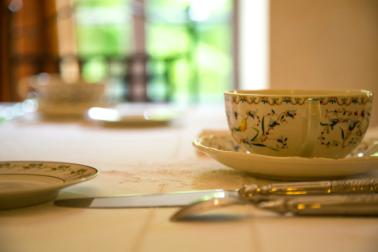 Table élégante dressée avec porcelaine décorée de motifs floraux bleus et or, couverts dorés, serviette blanche, et vue sur le jardin en arrière-plan au Château Valoux à Bordeaux.