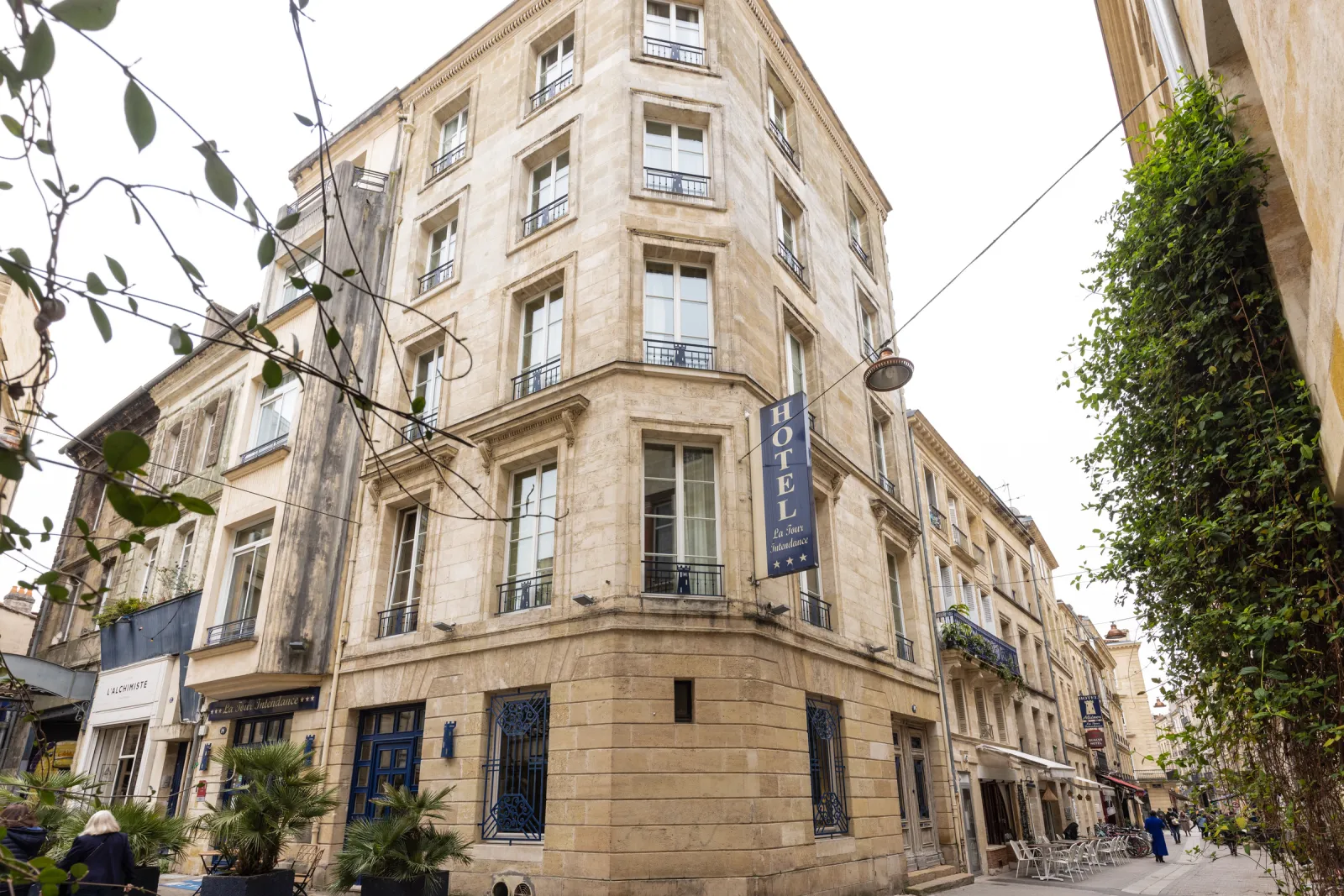 Façade de l'Hôtel de la Tour Intendance à Bordeaux, bâtiment historique en pierre de taille avec six étages, fenêtres à balcons, enseigne bleue d'hôtel, entouré de végétation et de façades anciennes.