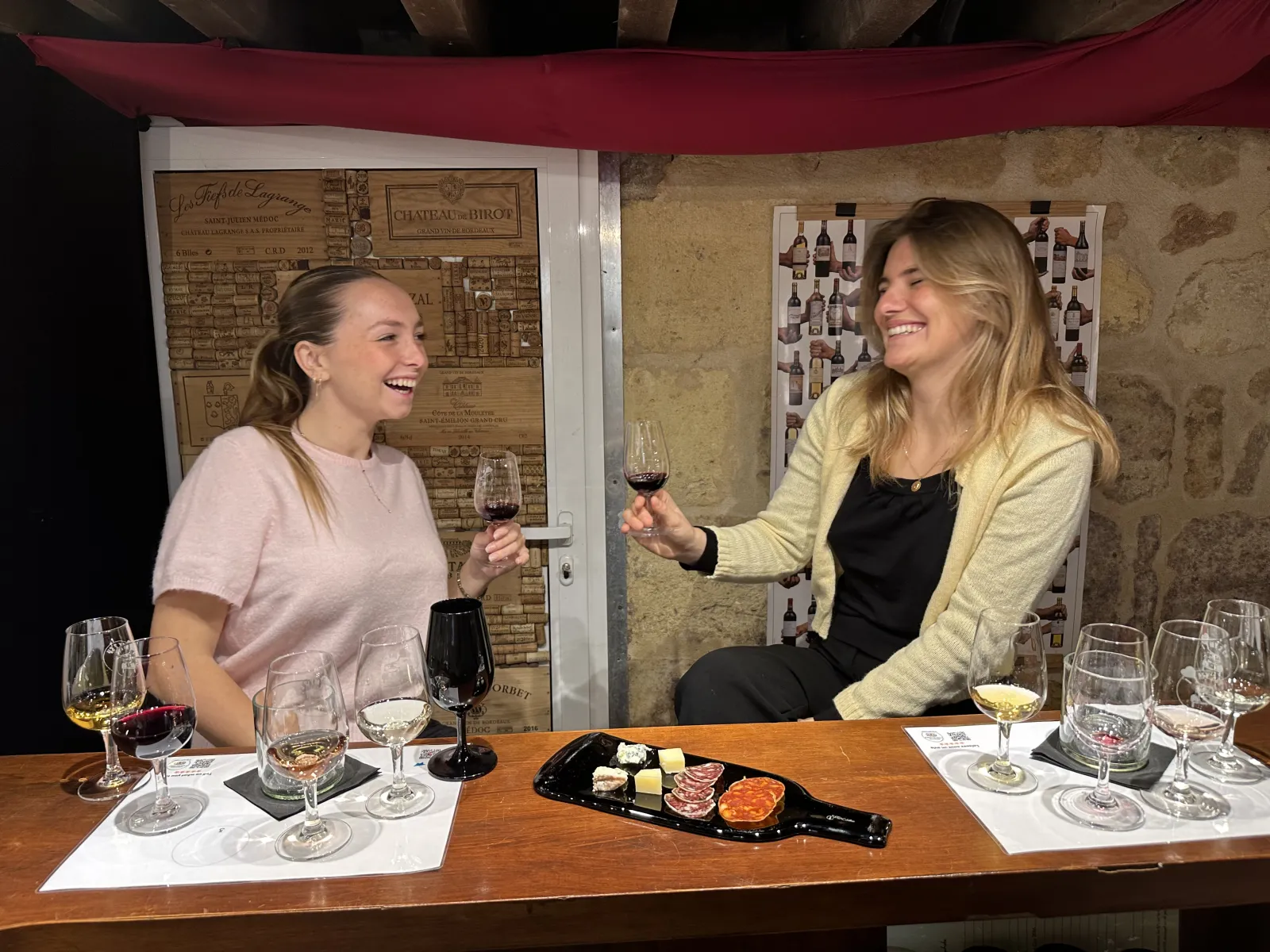 Deux femmes dégustent du vin rouge à Bordeaux lors d'une séance de dégustation. Elles sont assises à une table en bois avec des verres de vin, des verres vides numérotés et une assiette de charcuterie et fromage. Des affiches de vignobles sont visibles en