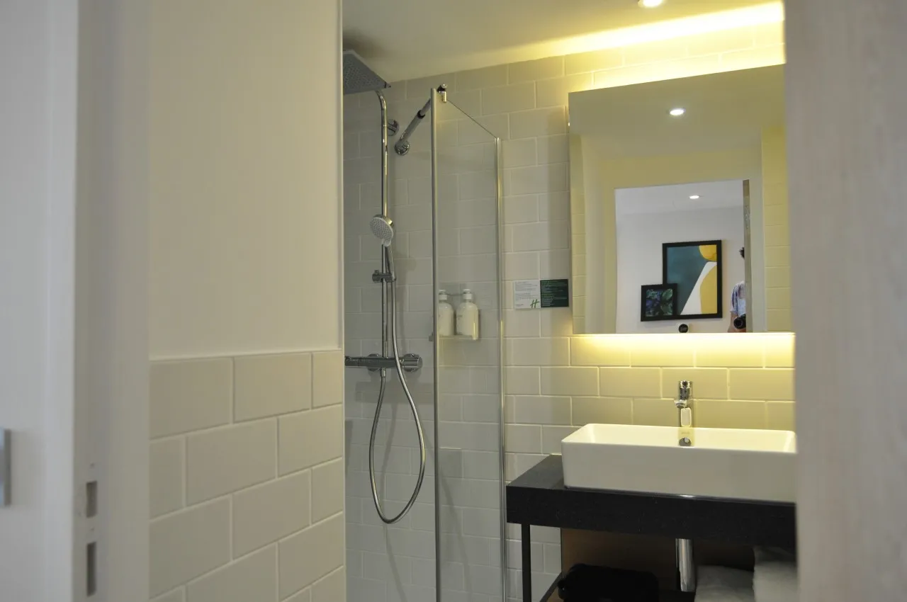 Salle de bain moderne de l'Holiday Inn Bordeaux Mérignac avec douche à l'italienne, vasque blanche sur meuble noir, miroir éclairé, carrelage blanc et éclairage ambiant jaune.