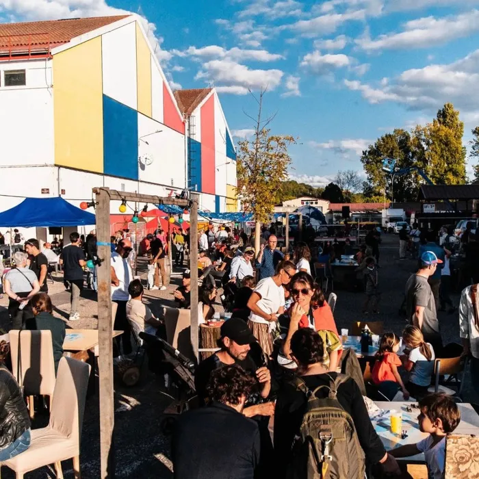 Foule festive lors de la 4ème édition de La Cité Bleue en fête à Bordeaux, avec un bâtiment coloré aux façades jaune, bleu et rouge, des tentes, des visiteurs et des food trucks sous un ciel bleu dégagé.