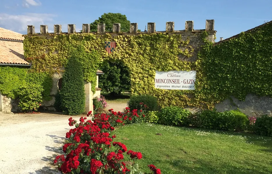 Entrée du Château Monconseil-Gazin à Bordeaux, avec ses murs de pierre couverts de lierre vert, un panneau d'identification, des roses rouges en première ligne et un jardin paysagé.