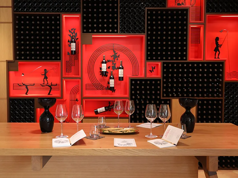 Présentation de dégustation de vins au Château Marquis d'Alesme Becker à Bordeaux, avec une table en bois, des verres de dégustation, des bouteilles de vin et un mur d'exposition rouge et noir décorés de motifs et de silhouettes.