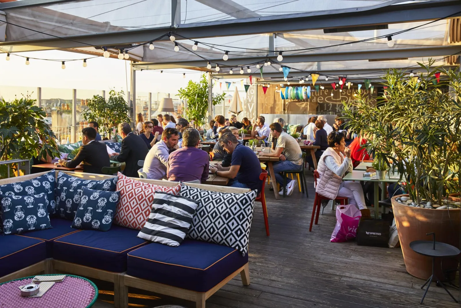 Terrasse du restaurant Mama Shelter à Bordeaux avec clientèle assise sous une toiture rétractable, décorée de guirlandes lumineuses, de coussins colorés et de plantes vertes, offrant une vue sur la ville.