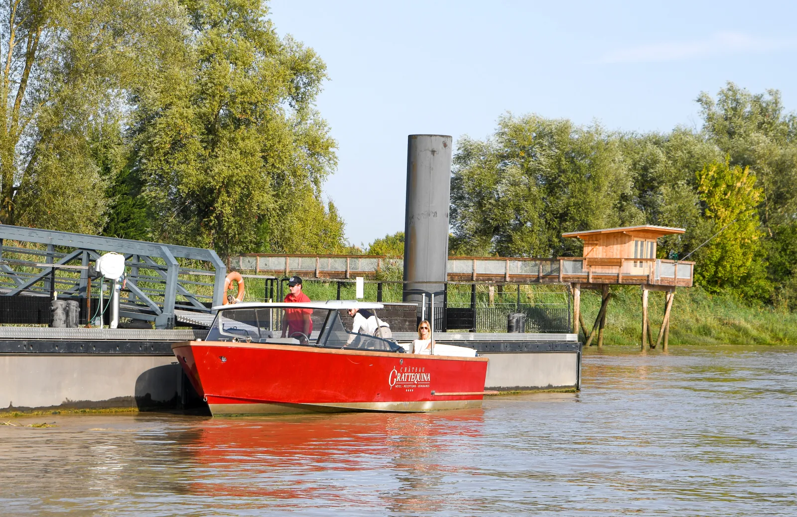 Le bateau taxi rouge Grattequina navigue sur la Garonne à Bordeaux avec des passagers à bord, devant des installations portuaires et des arbres.