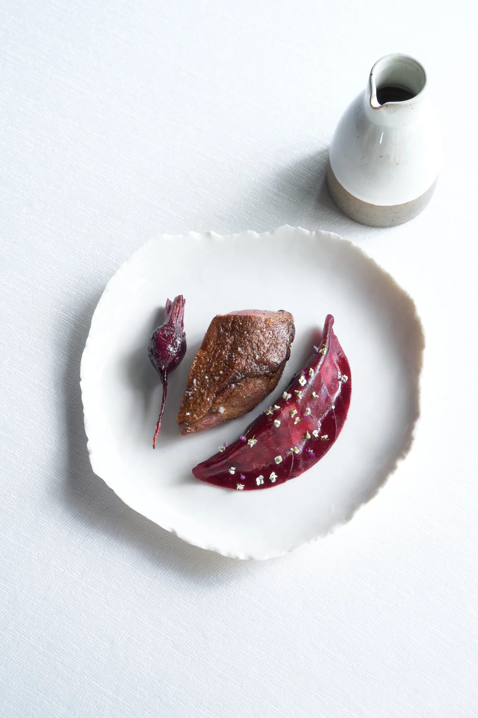 Assiette blanche avec un filet de viande cuit accompagné de betteraves décorées et d'une sauce, avec une petite carafe blanche en arrière-plan