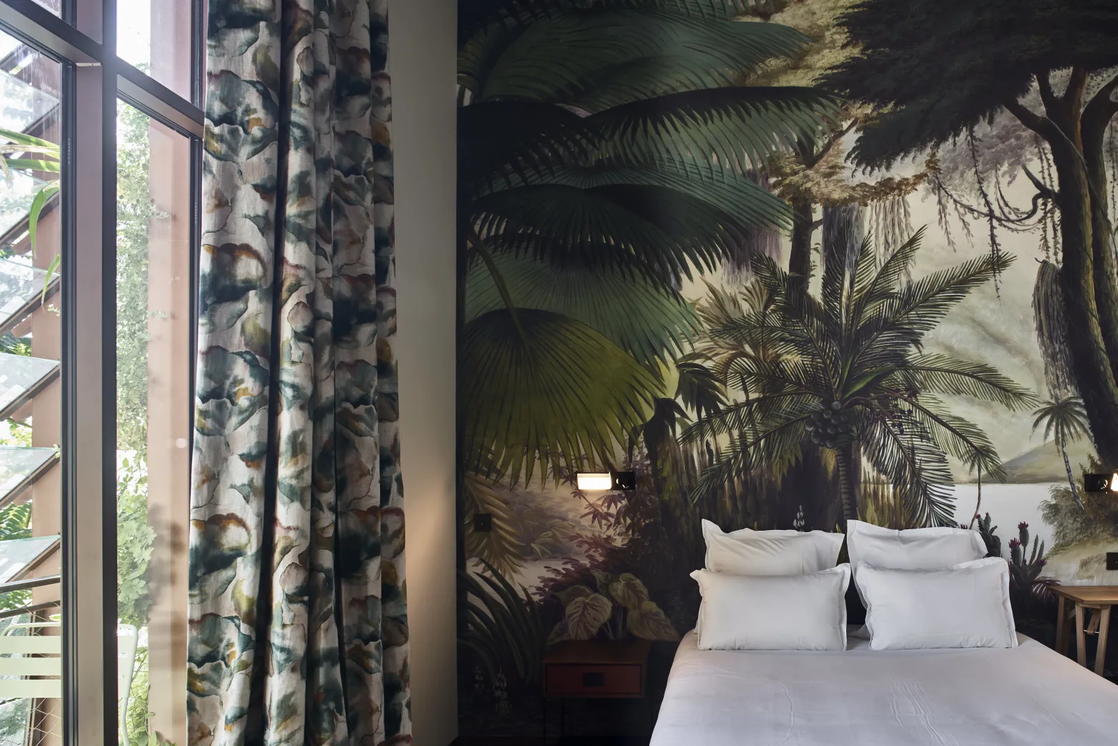 Chambre de l'Hôtel La Zoologie à Bordeaux avec un lit blanc, une fresque murale représentant une forêt tropicale avec des palmiers, des rideaux à motifs floraux et une baie vitrée donnant sur la verdure extérieure.