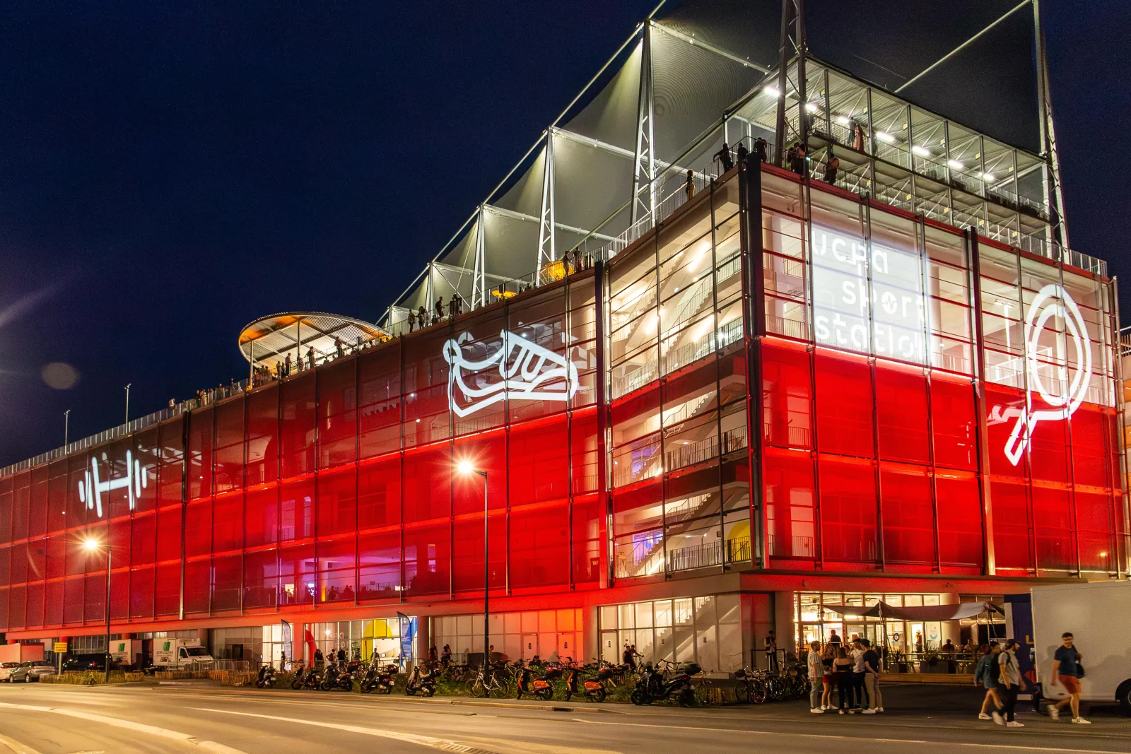 Façade illuminée en rouge de la UCPA Sport Station à Bordeaux, avec sa structure architecturale moderne en verre et acier, photographiée de nuit avec des visiteurs devant l'entrée.
