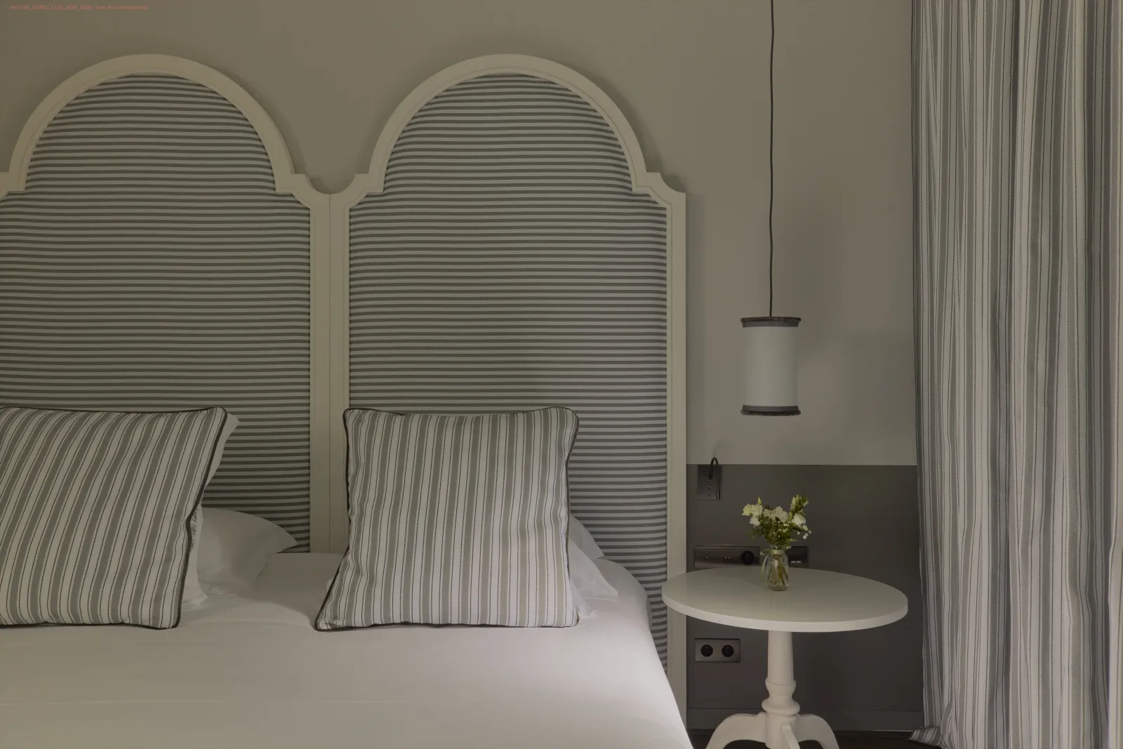 Chambre élégante avec lit double aux têtes de lit arquées rayées, oreillers assortis, table de nuit ronde blanche avec vase de fleurs, rideaux striés et lampe suspendue.