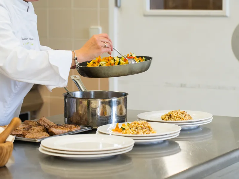 Chef en veste blanche versant des légumes sautés d'une poêle vers une casserole lors d'un atelier culinaire au Château Kirwan à Bordeaux, avec des assiettes dressées de maïs et légumes sur le plan de travail.