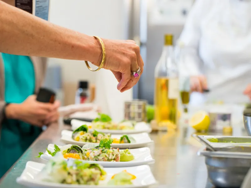 Atelier de cuisine au Château Kirwan à Bordeaux : une personne en tenue turquoise prépare des plats gastronomiques garnis d'herbes fraîches et de citron, avec des bouteilles d'huile d'olive en arrière-plan.