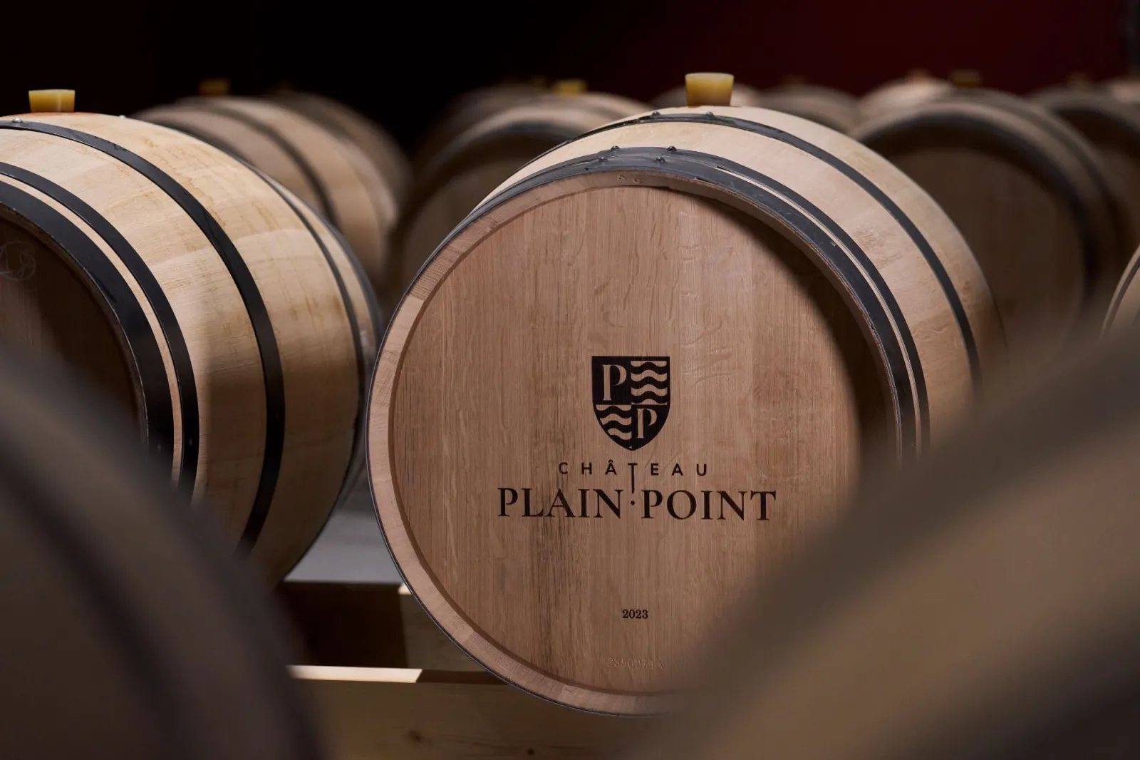 Rangée de tonneaux en bois portant le logo et le nom du Château Plain Point, datés de 2023, dans une cave de vieillissement de vin à Bordeaux.