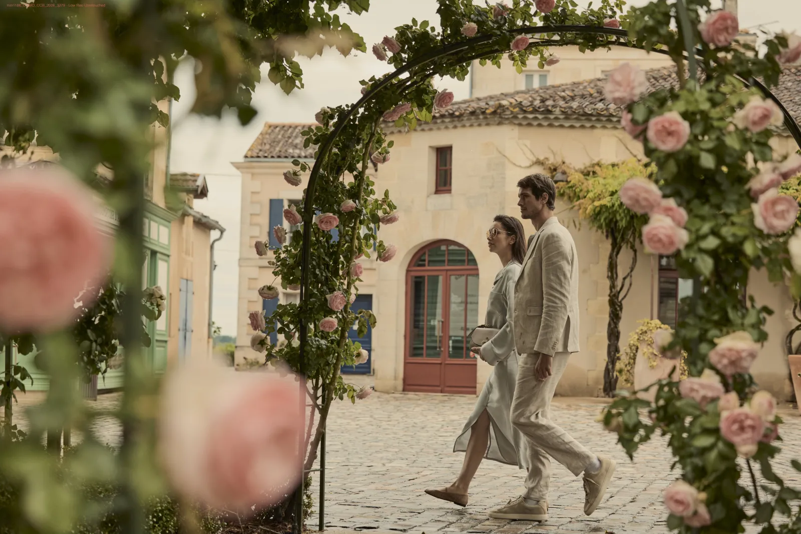 Couple marchant dans une cour pavée bordée de roses roses grimpantes, devant des bâtiments en pierre de style méditerranéen avec portes rouges et fenêtres bleues.