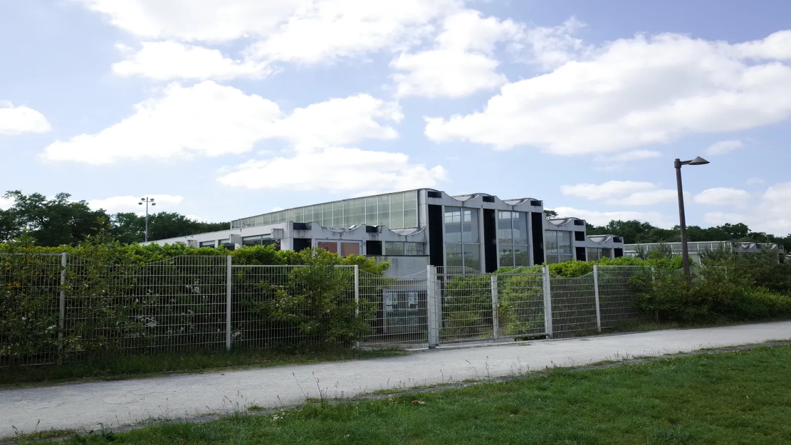 Le Stade nautique Henri Deschamps de Bordeaux, bâtiment moderne en verre et métal avec toiture en dents de scie, entouré d'une clôture métallique et de végétation, sous un ciel nuageux.