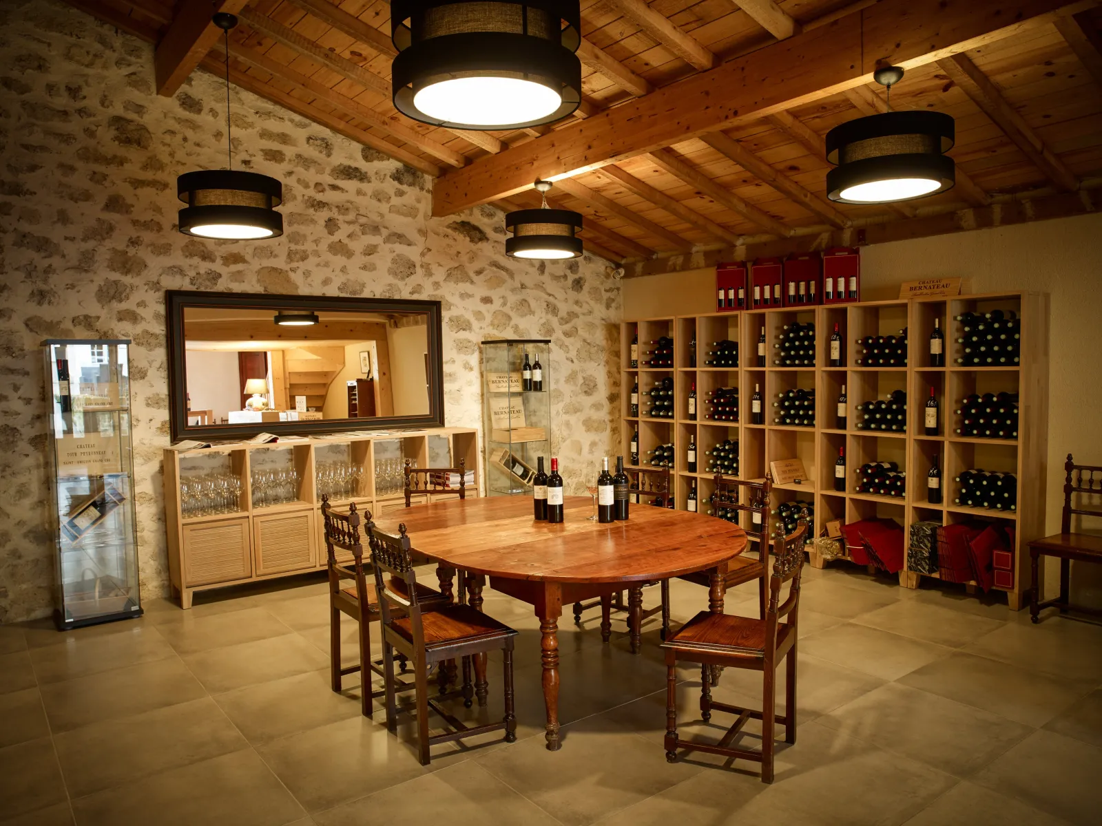 Salle de dégustation du Château Bernateau à Bordeaux avec table ronde en bois, chaises traditionnelles, étagères de bouteilles de vin, comptoir de service et décoration rustique avec poutres apparentes.