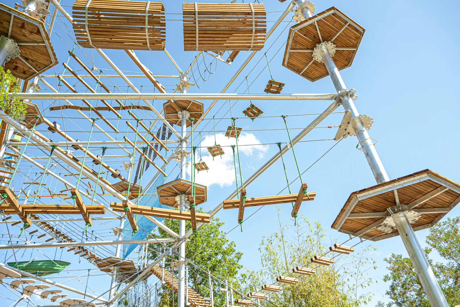 Vue aérienne d'une structure de parc d'aventure en bois avec des parcours suspendus, des poutres métalliques et des éléments de jeu aériens entrelacés de cordes et de câbles, sous un ciel bleu dégagé.