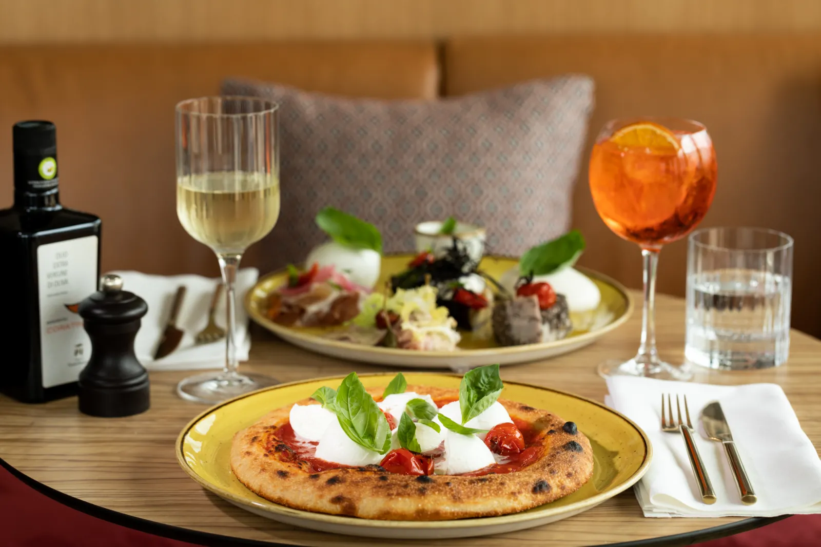 Table de restaurant élégante avec deux pizzas garnies de mozzarella et tomates, accompagnées de verres de vin blanc et d'un cocktail orange, dans une ambiance raffinée de Bordeaux.