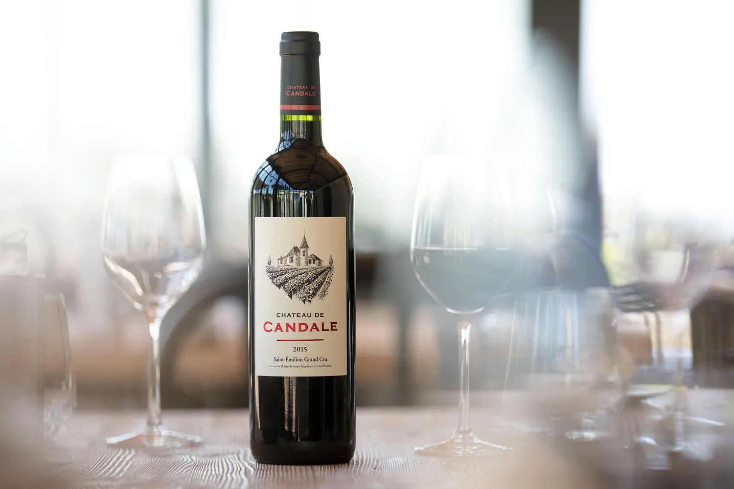 Bouteille de vin rouge Château de Candale 2015, Saint-Émilion Grand Cru, entourée de verres à vin vides sur une table blanche avec un arrière-plan flou de Bordeaux.